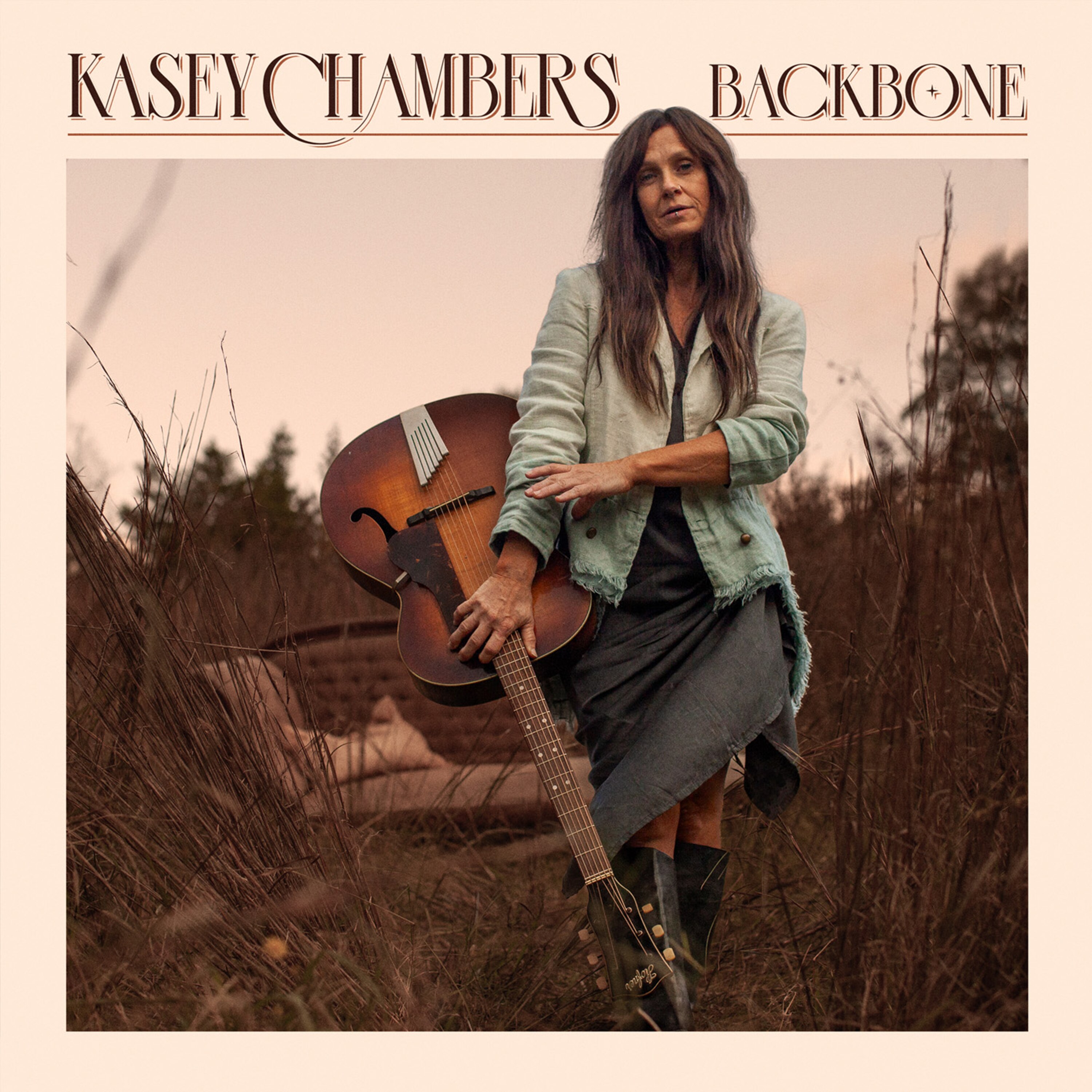 Kasey Chambers stands in white jacket with a guitar by a couch chair in a field of reeds. Text reads: Kasey Chambers - Backbone