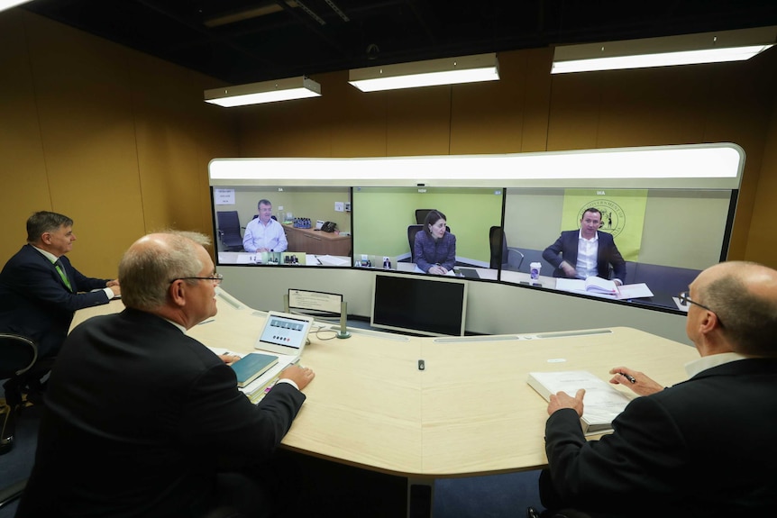 Three men sit at a semi circle table in suits in front of three large screens with one person in each.