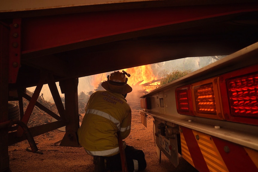 A firefighter crouches in front of a bushfire.