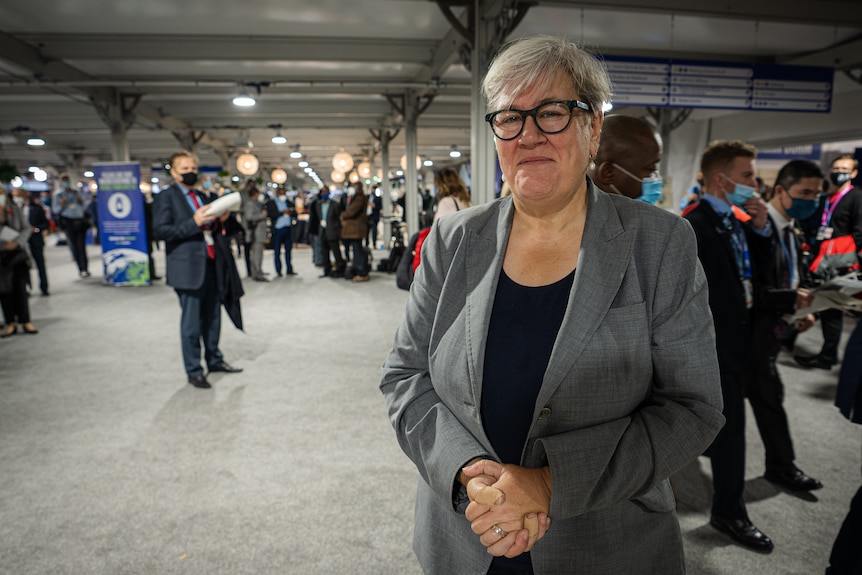 A woman in a grey suit holds her hands in a conference centre.