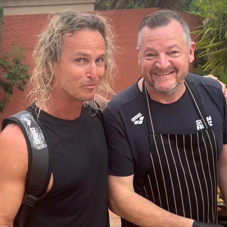 man with long curly blonde hair in black singlet and backpack with grey hair and bearded man in striped apron