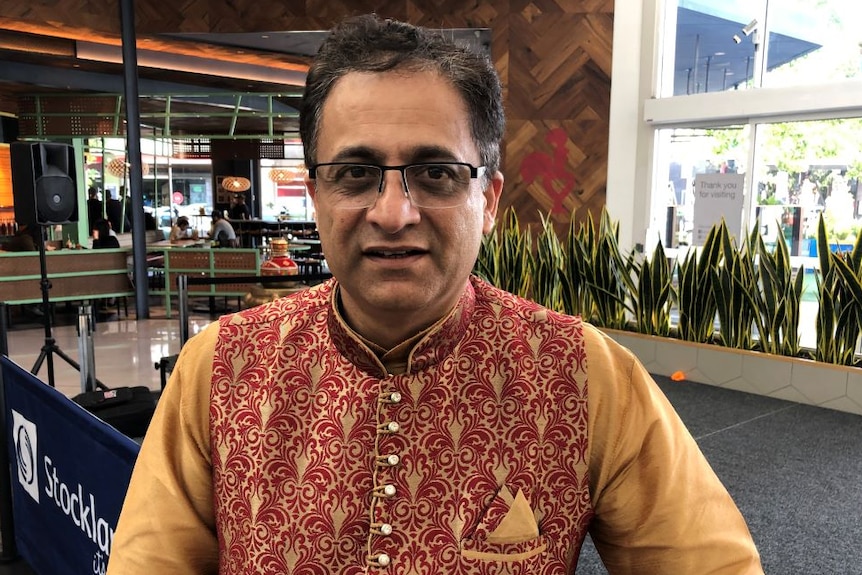 A man with glasses, wearing a red and gold Indian shirt stands in a shopping centre.