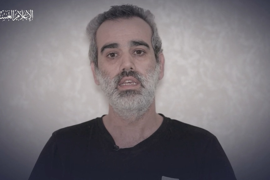 A man with dark eyebrows and a grey beard in a black t shirt in front of a grey background