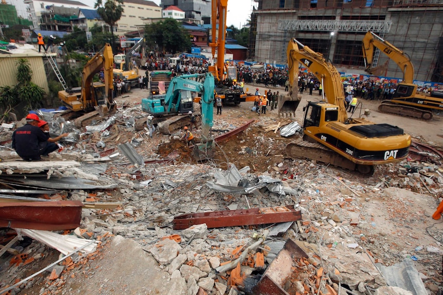 A wide photo looks down on the debris of a collapsed building site, with five excavators and a large crowd surrounding it.