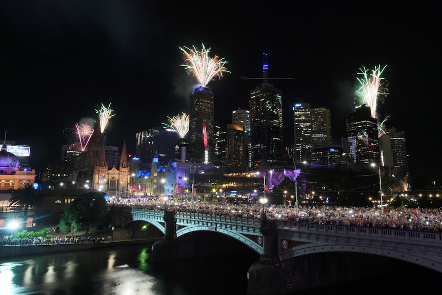 Fireworks explode over several buildings in Melbourne's CBD. Thousands of people stand along a bridge.