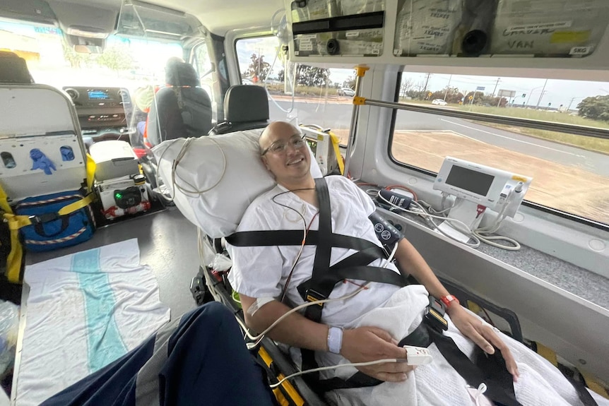 A man smiles at the camera while strapped to a gurney in an ambulance 