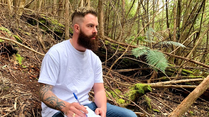 A bearded young man sits writing in the bush