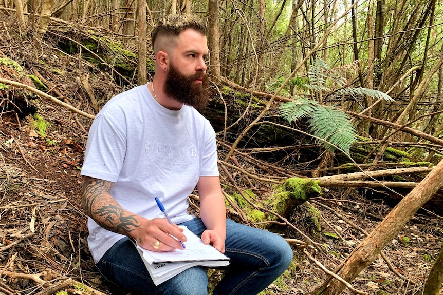 A bearded young man sits writing in the bush