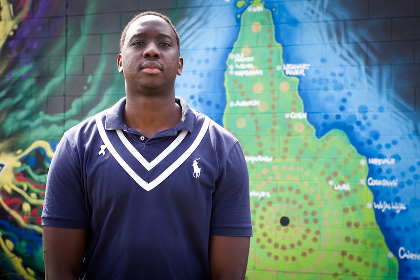 An Aboriginal man standing in front of a brightly coloured painted map of far north Queensland.