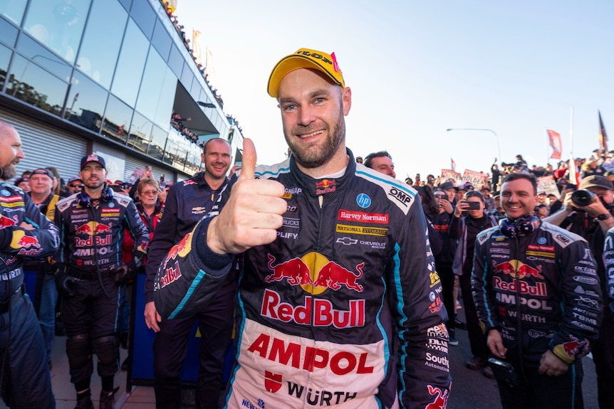 Race driver standing in front of his team with his thumb up.