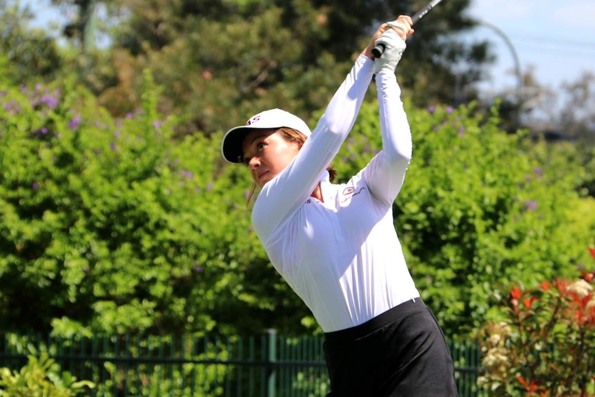 Becky Kay, dressed in a white shirt and white cap, swings a golf club