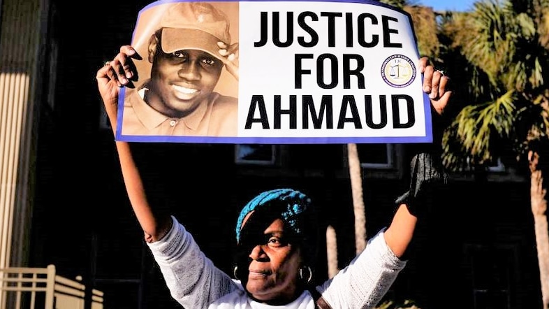 A woman holds up a sign that reads justice for Ahmaud out the front of a courthouse on a bright blue sky day 