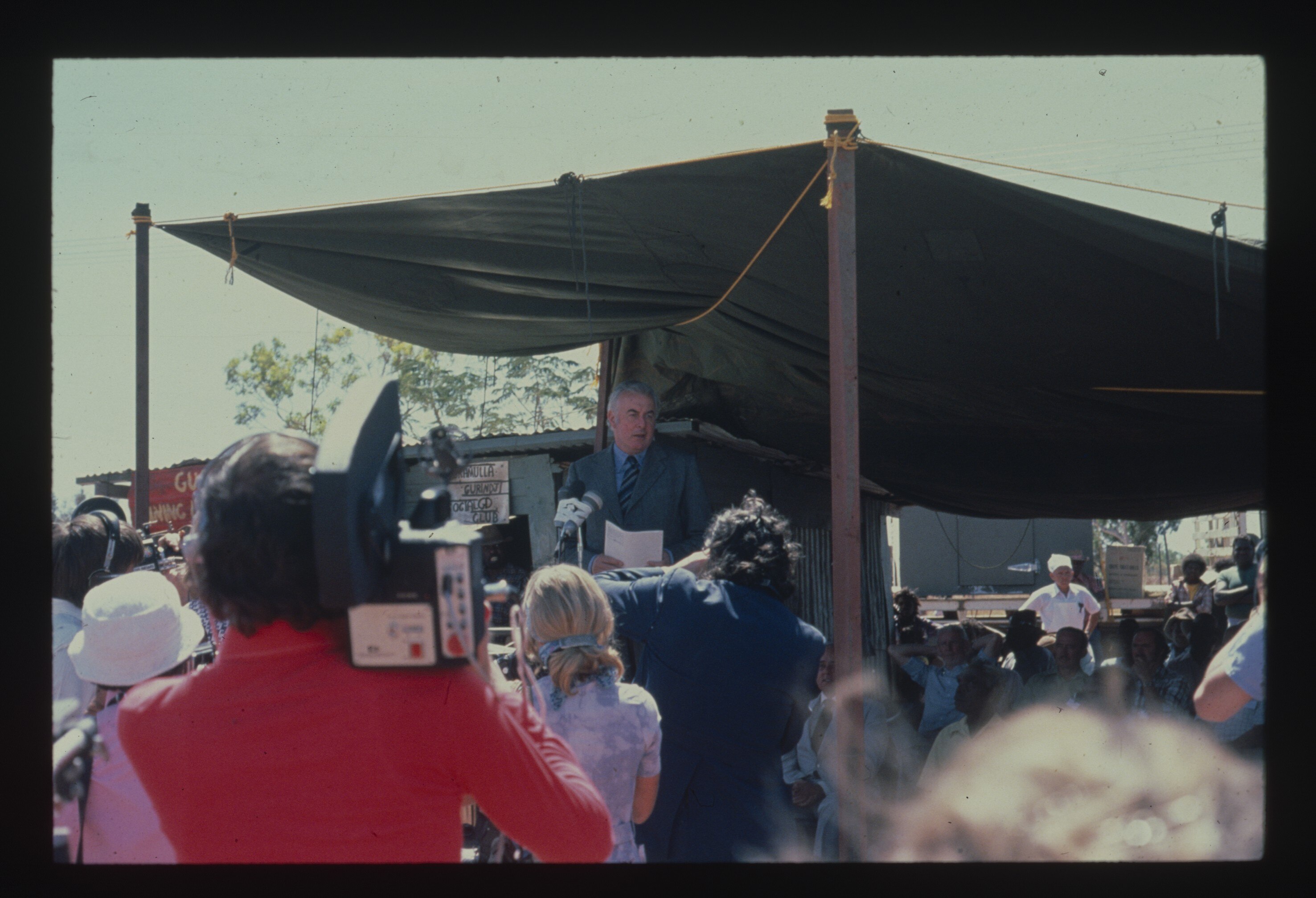 A man talking at a podium some distance away, white crowd of camera operators filming in front. 