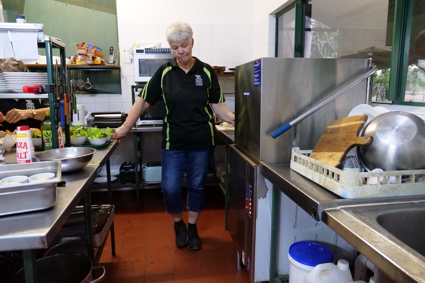 A woman with a black t-shirt and grey hair looks down while standing in the middle of a kitchen. 