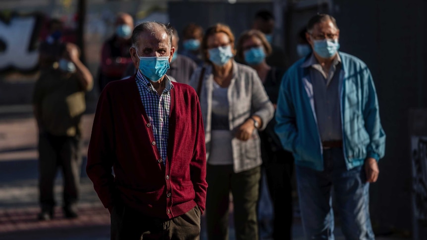 Elderly people wearing disposable face masks stand in a socially-distant queue in dappled sunlight