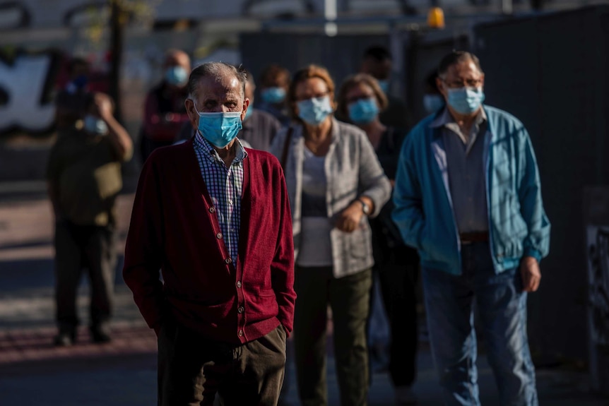 Elderly people wearing disposable face masks stand in a socially-distant queue in dappled sunlight