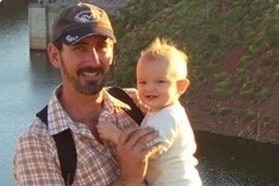 A man with a beard stands in front of a lake, holding a baby.