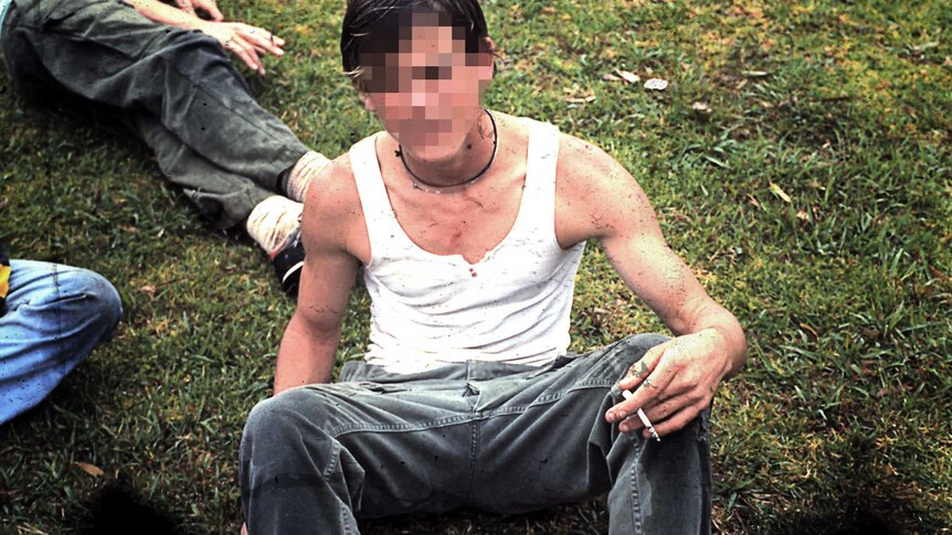 A teenage boy sits in a field smoking a cigarette.