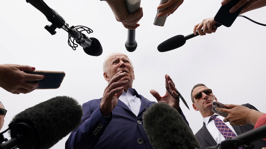 An elderly man and a man wearing sunglasses stand in front of people holding microphones