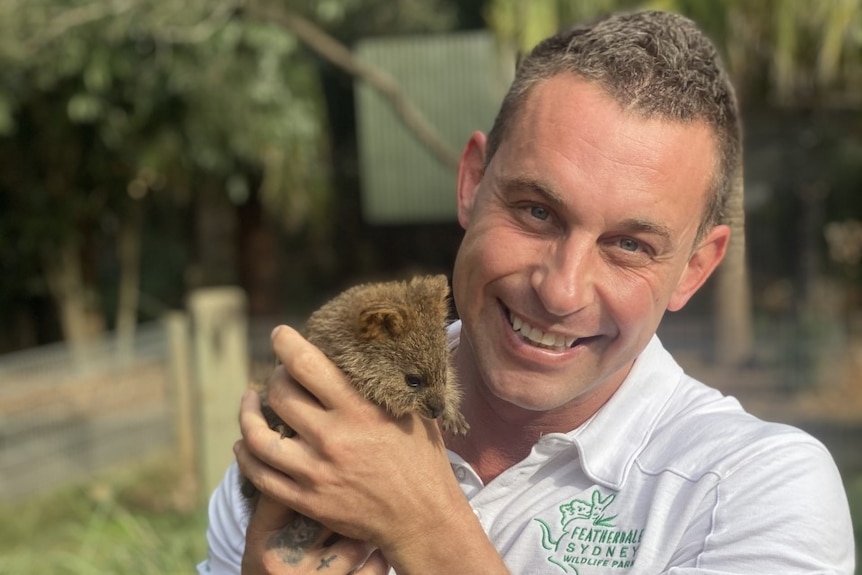 A man wearing a white polo shirt holds a small mammal up to his face while smiling at the camera.