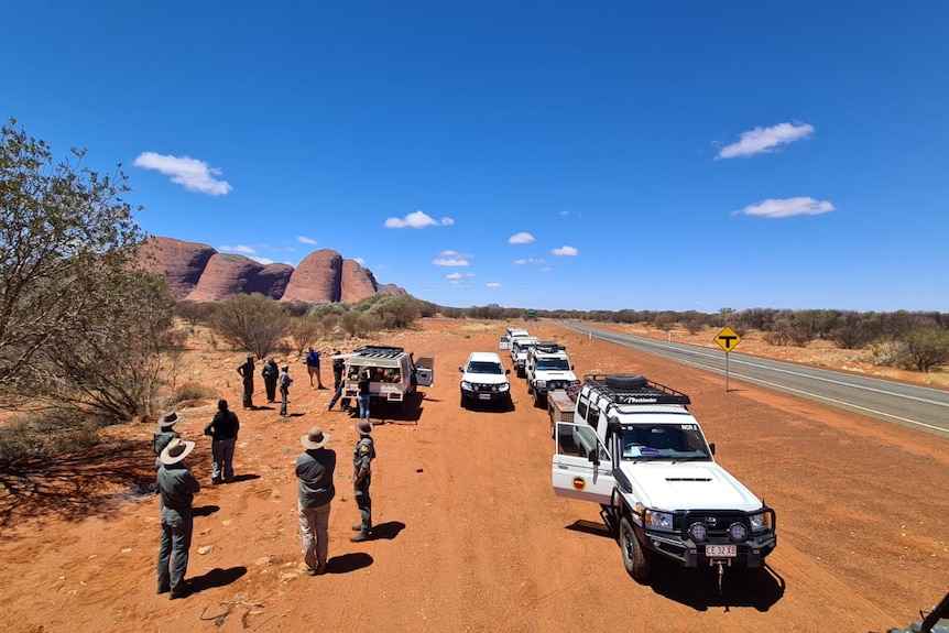 A shot from the air of a convoy of four wheel drives an about ten people standing by the side of an outback road.