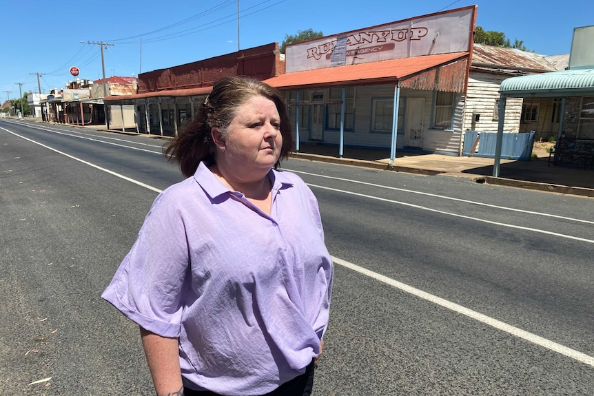 A woman stands looking down a deserted street of a small country town. There are some disused shops behind her.