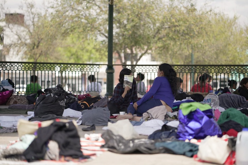 A woman wearing a purple shirt talks to another woman holding a book surounded by a pile of bags.