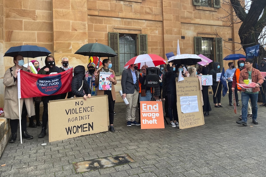 People stand holding umbrellas and signs with writing against wage theft and violence against women