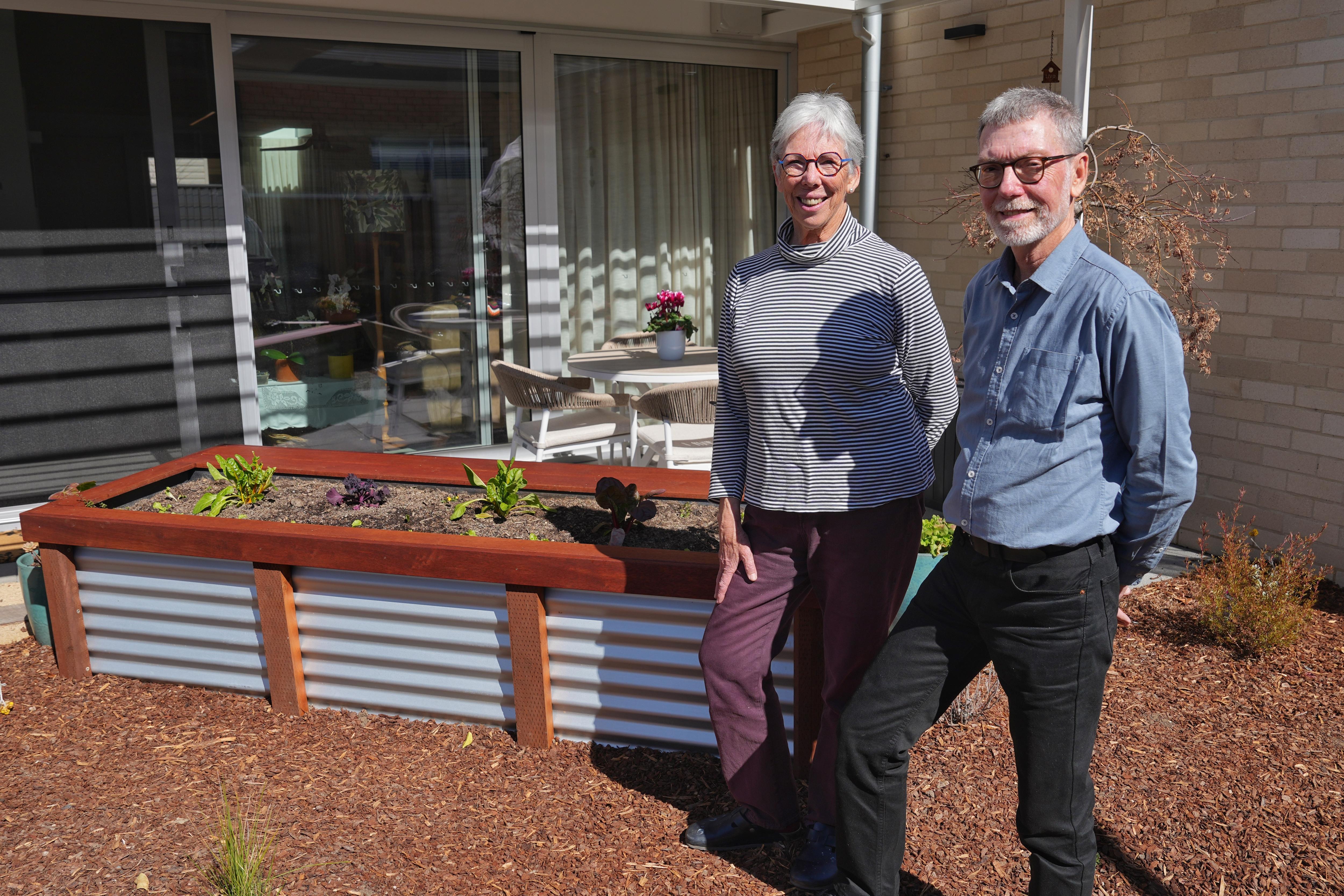 Trish and Ian, two older people, stand and smile in a garden outside a house.