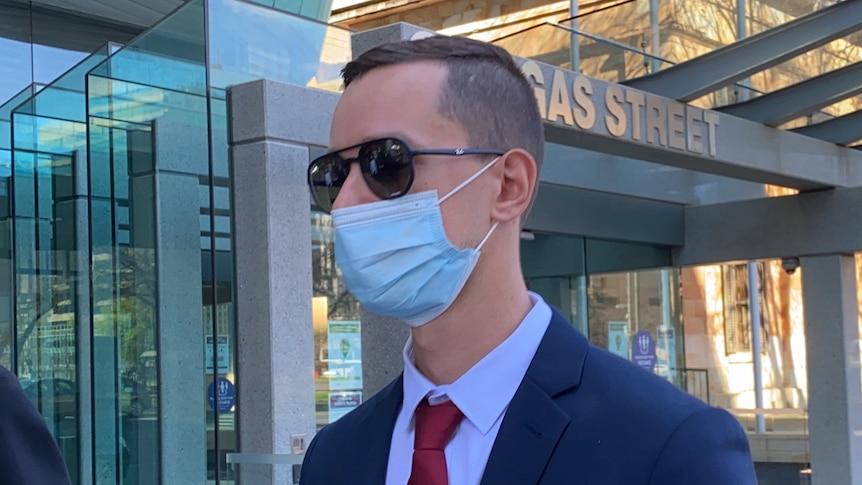 A man wearing a face mask, sunglasses and a blue suit outside a court building