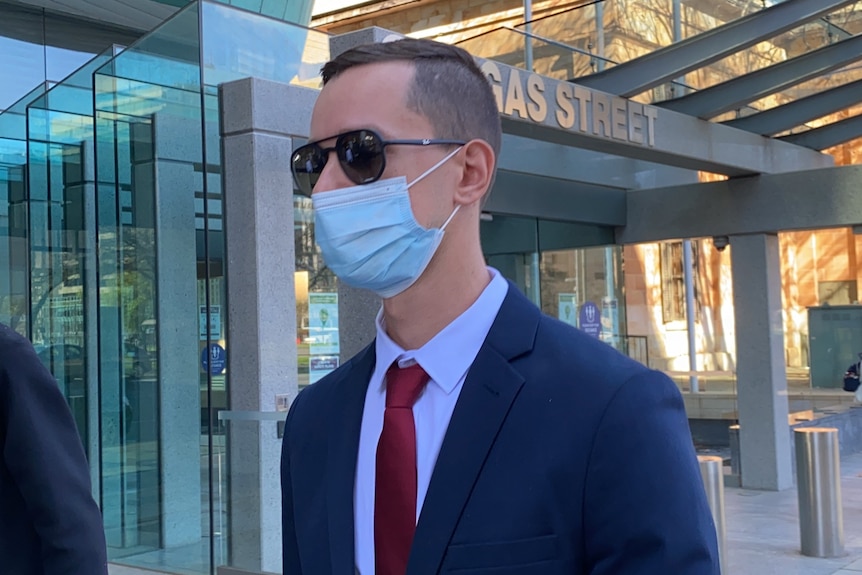 A man wearing a face mask, sunglasses and a blue suit outside a court building