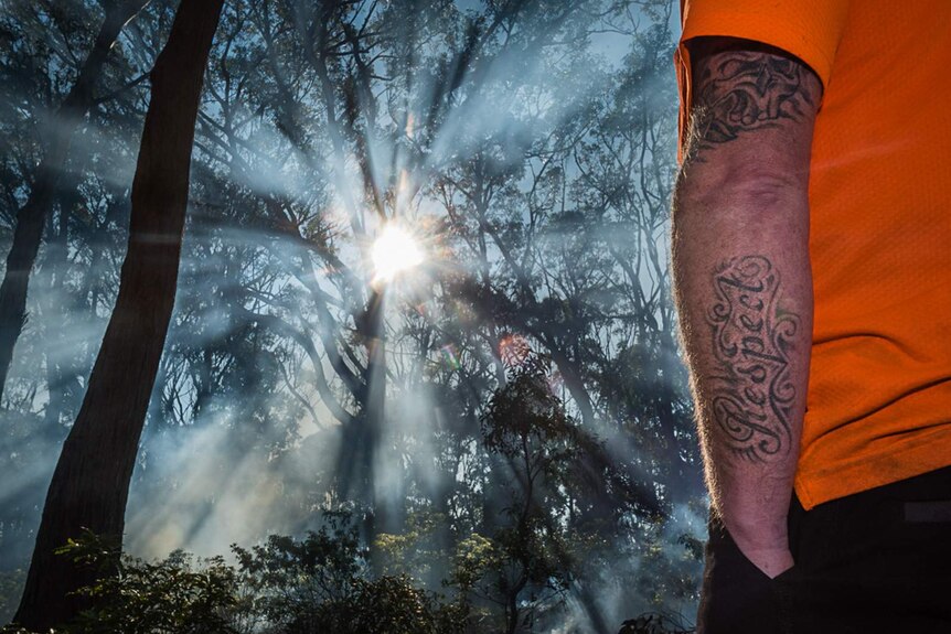 Smoke from cultural burn coming through trees, rear of man with arm in pocket with a 'Respect' tattoo in foreground