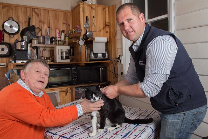 A man sitting at a table holding with a cat on the table and another man standing and both are holding the cat