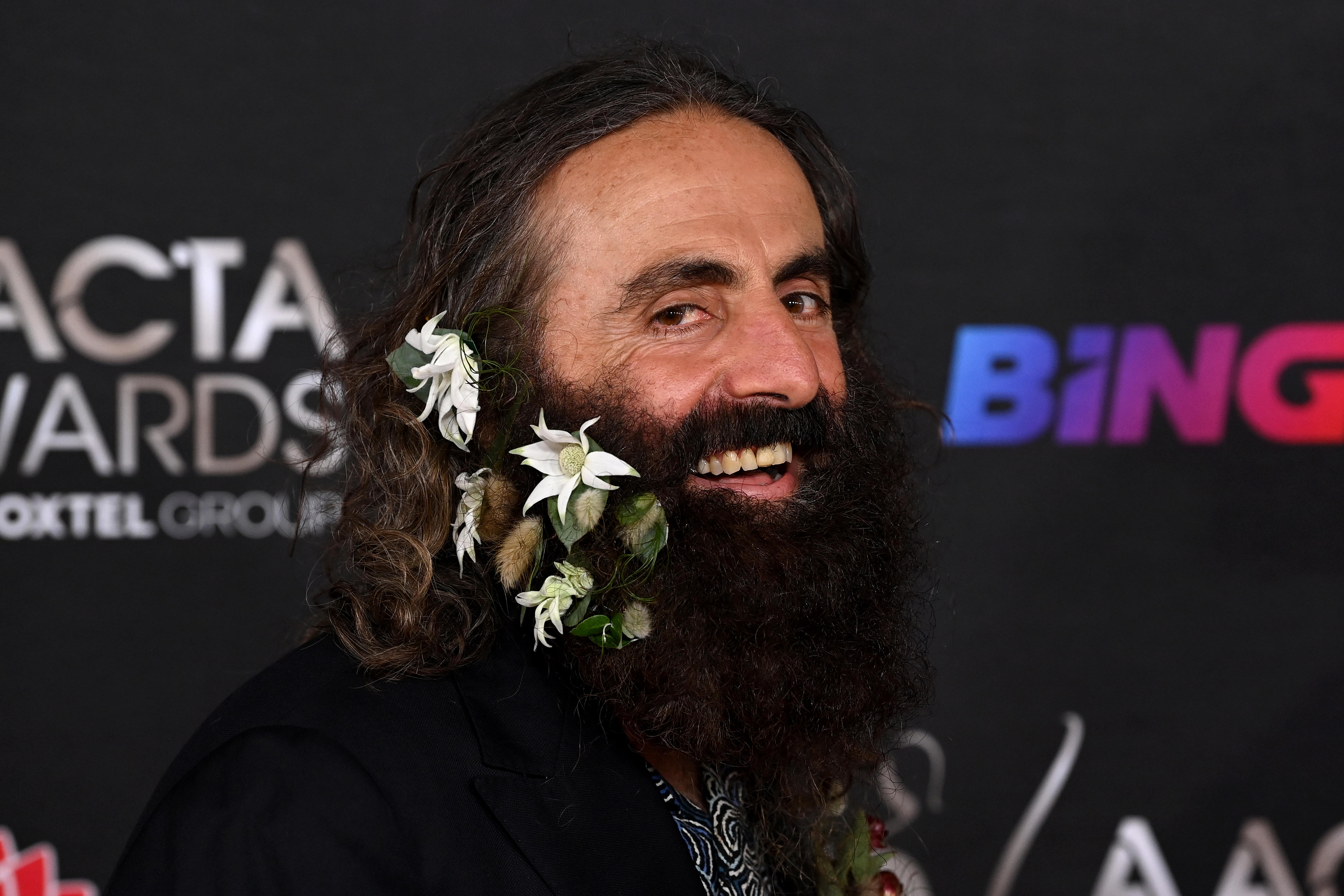 A man with a thick dark beard and flowers adorning his facial hair smiles at the camera 