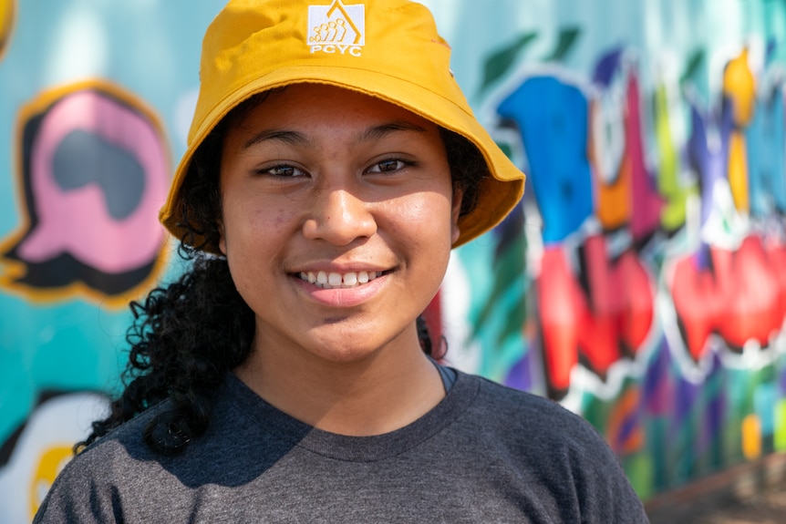 A young girl with curly black hair and a yellow cap smiles.