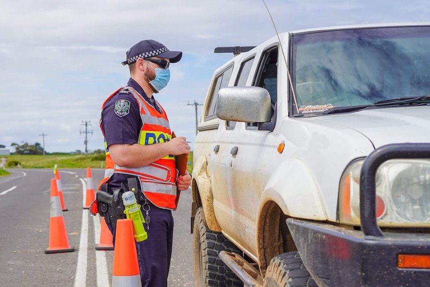 A police officer in a high vis vest and face mask talks to a man through a car passenger door.