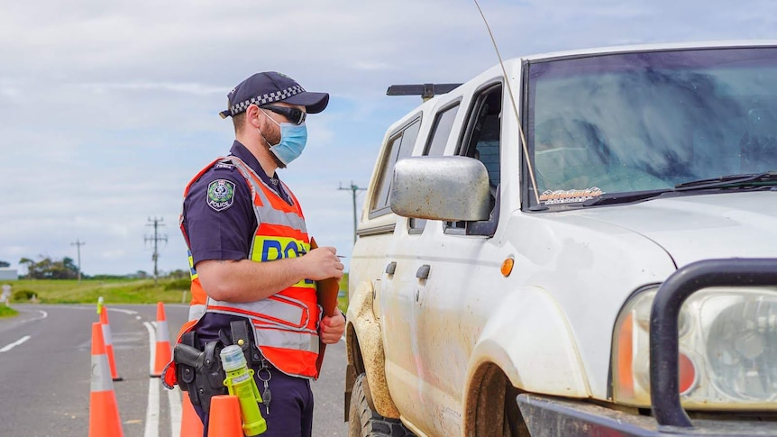 A police officer in a high vis vest and face mask talks to a man through a car passenger door.