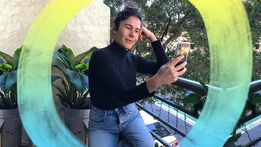 A woman sits on a balcony looking into her phone and smiling in a story about what makes a good, happy life.