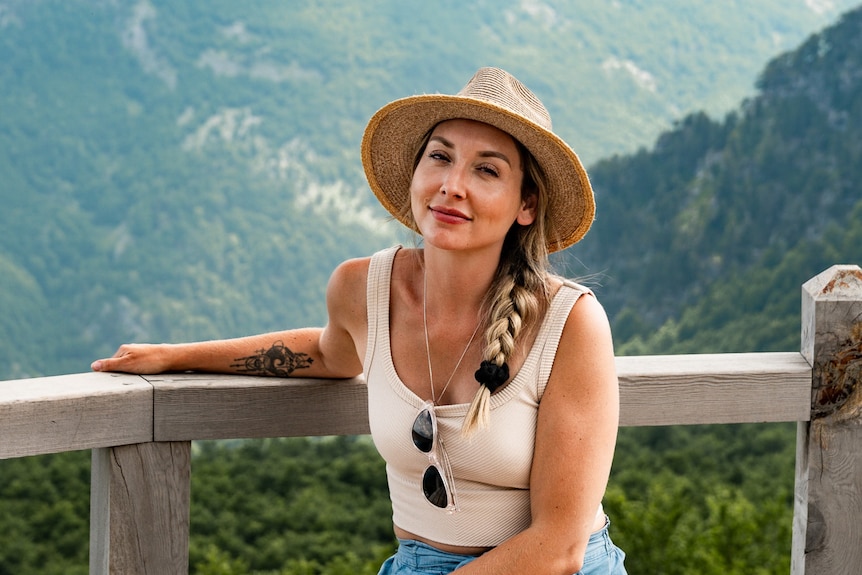 A blonde woman smiling while posing near a mountain wearing a hat.