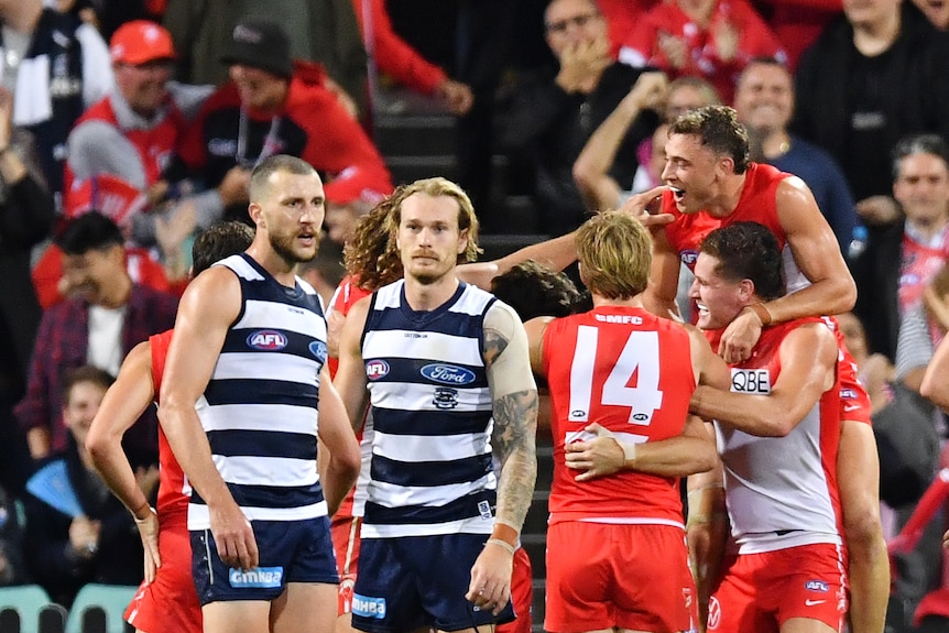 Winning AFL players leap into each others arms at the end of a game as the losing side look dejected.