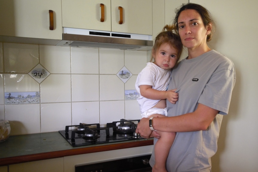A lady standing in a kitchen holden a small child next to a stove