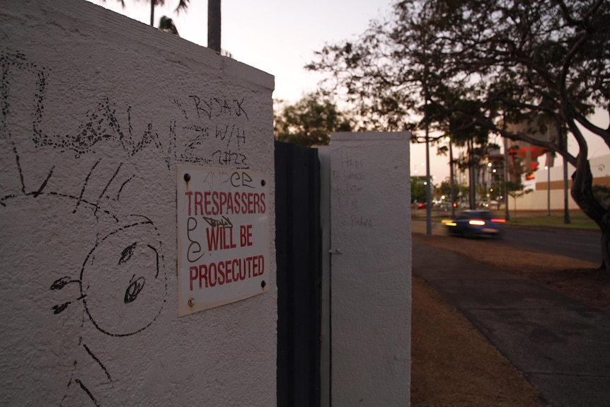 A sign on a wall with graffiti that says 'Trespassers will be prosecuted'. It is dusk and a car is driving by.