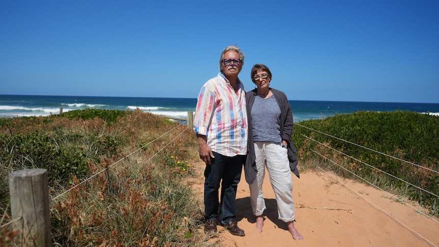 a man and a woman, both wearing glasses, stand on a beach on the grassy dunes