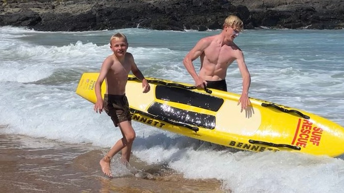 two blonde teenagers in surf holding life saving paddle board