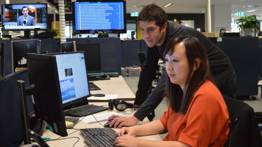 Wide shot of Viney and Zhou looking at computer screens in newsroom.