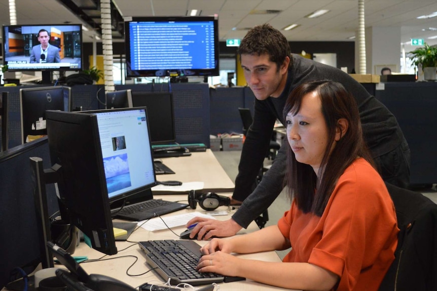 Wide shot of Viney and Zhou looking at computer screens in newsroom.