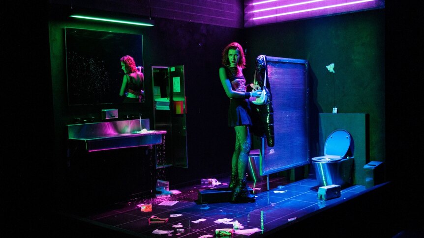 White trans woman with red hair wears pink corset, black skirt and tights in pink and green neon-lit bathroom.