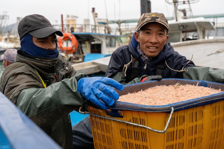 A close up of two men wearing caps and large jackets holding a bucket of fish.