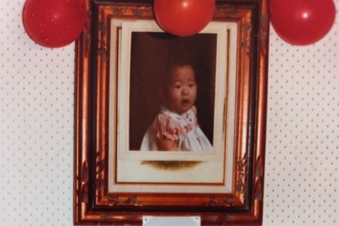 A portrait of a baby is seen on a wall with a Korean flag under it and a birthday cake on a table below that.