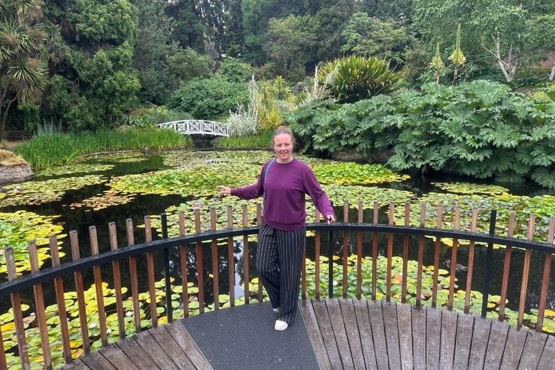 A smiling woman leans against a railing in front of a pond dotted with lily pads.